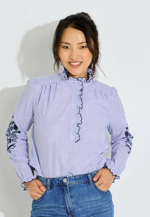 Mujer sonriente que lleva una blusa de rayas azules y blancas con cuello y puños con volantes, bordado floral en las mangas y vaqueros azules.
