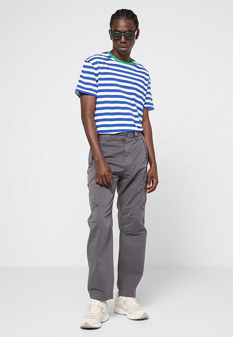 T-shirt à rayures bleu et blanc à manches courtes avec encolure verte, associé à un pantalon cargo gris et des baskets blanches, comportant plusieurs poches.