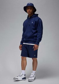 Marineblå hættetrøje med frontlomme, marineblå shorts, hvide sokker og hvide sneakers. Alle genstande har Jordan-logoet. En bøttehat fuldender outfittet.