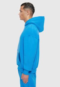 Niebieska hoodie wykonana z gładkiego materiału, z kieszenią z przodu i ściągaczami przy mankietach. Pokazana z boku, podkreślająca swój luźny krój.
