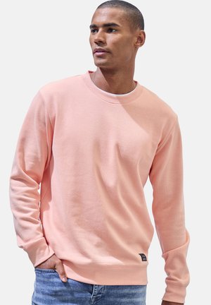 Mann trägt hellrosa Rundhalsausschnitt-Sweatshirt und blaue Jeans, steht mit einer Hand in der Tasche und schaut zur Seite.