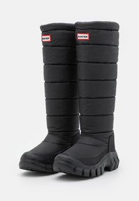 Svart, vattentäta knähöga stövlar med en vadderad överdel, texturerad yta och kraftig gummisula. Har röd och vit branding på skaftet.