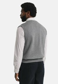 Grijze wollen vest met een geribde zoom, gedragen over een wit en rood gestreept overhemd. De afbeelding toont de achterkant van de outfit.