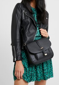 Svart läder crossbody-väska med klaffstängning och guldaccent. Bärs med en grön leopardmönstrad klänning och svart läderjacka.