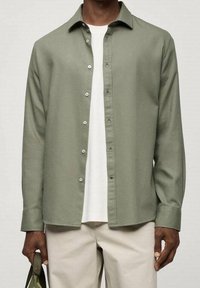 Camicia button-up verde chiaro in tessuto testurizzato, con colletto e pattina frontale, indossata sopra una t-shirt bianca, abbinata a pantaloni chiari.