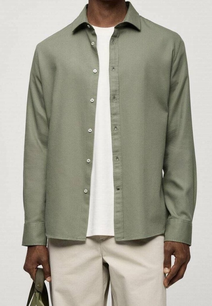 Camicia button-up verde chiaro in tessuto testurizzato, con colletto e pattina frontale, indossata sopra una t-shirt bianca, abbinata a pantaloni chiari.