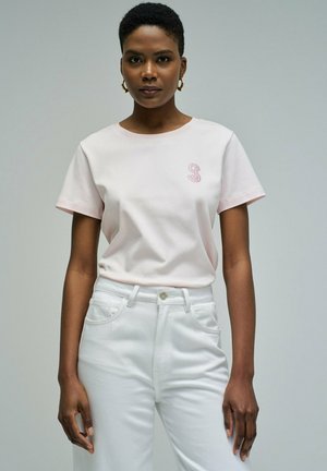 Mujer con cabello corto, vestida con una camiseta rosa claro con un pequeño emblema bordado y pantalones blancos de cintura alta, de pie frente a un fondo liso.