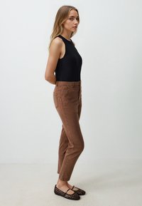 Top nero senza maniche, pantaloni marroni aderenti con tasche e ballerine intrecciate nere. Texture liscia, silhouette slim e colori delicati.