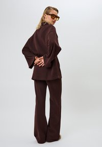 Haut en tricot côtelé marron avec de larges manches et un pantalon évasé assorti. Complété par des lunettes de soleil. Modèle tourné de dos par rapport à la caméra.