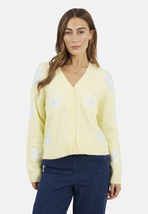 Kobieta ubrana w jasnożółty sweter z wzorem białych i jasnoniebieskich stokrotek, zapinany z przodu na guziki, w połączeniu z ciemnoniebieskimi dżinsami.