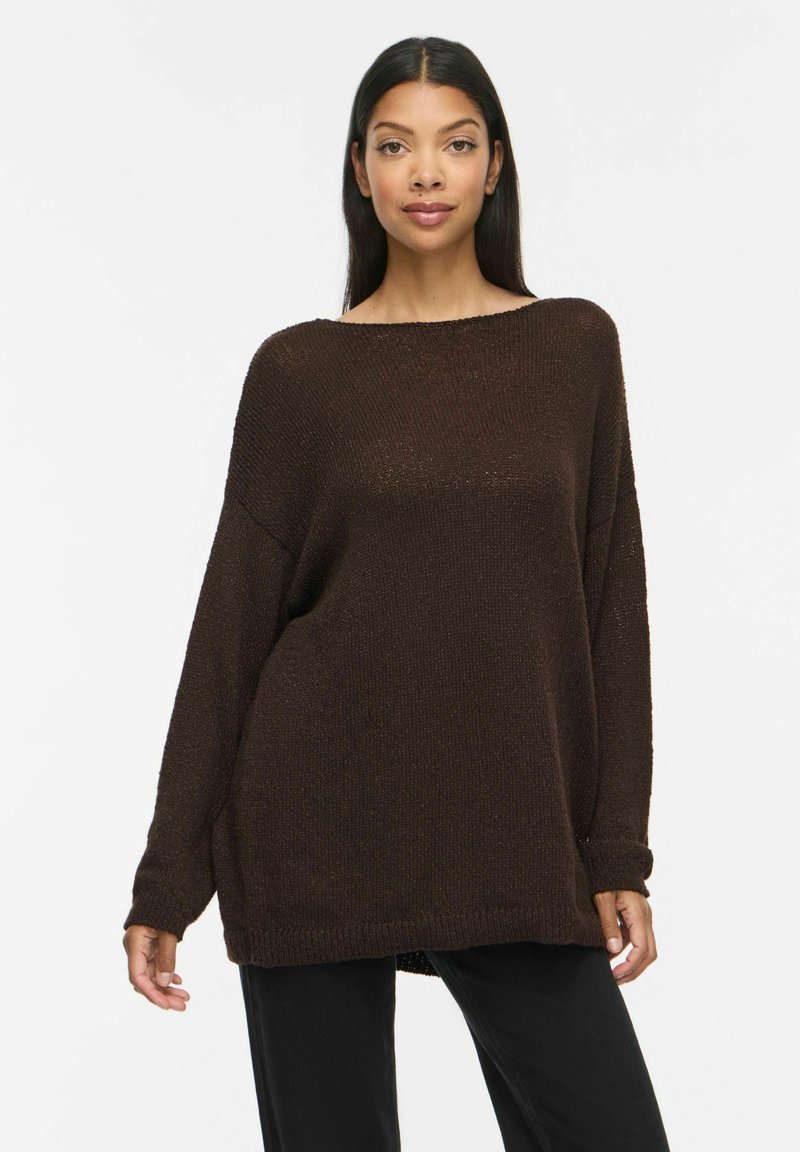 Femme aux longs cheveux foncés portant un pull en tricot marron foncé ample et un pantalon noir, debout devant un fond clair uni.