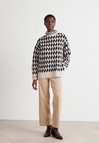 Übergroßer Pullover mit einem geometrischen Muster in Schwarz und Creme, gerippter Kragen und Saum. Kombiniert mit beigen, weit geschnittenen Hosen und dunklen Knöchelstiefeln.