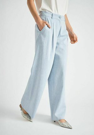 Personne portant un pantalon large bleu clair avec des poches latérales et des talons pointus blancs ornés de clous noirs, se tenant devant un fond uni.