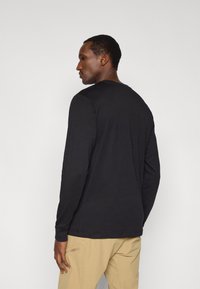 Μαύρο πουκάμισο με μακριά μανίκια, στρογγυλή λαιμόκοψη, εφαρμοστή γραμμή, απαλό ύφασμα, συνδυασμένο με μπεζ παντελόνι. Βλέπεται από πίσω, τονίζοντας το σχέδιο του πουκαμίσου.