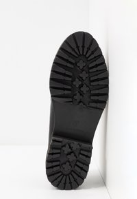 Semelle de botte en caoutchouc noir avec un motif profond et robuste de crampons comportant des formes en croix et des lignes pour l'adhérence, présentée sur un fond blanc.