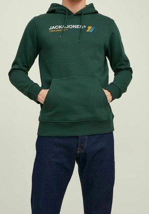 Muž nosící tmavě zelenou mikinu Jack & Jones s přední kapsou a modré džíny, ruce zasunuté v kapse mikiny, neutrální pozadí.
