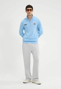 Sudadera azul claro con media cremallera y un pequeño logo, combinada con pantalones de chándal grises. El conjunto tiene un corte relajado y se acompaña de zapatillas deportivas blancas.