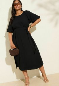 Schwarzes Midikleid mit kurzen Puffärmeln, tailliertem Bund und fließender Silhouette. Kombiniert mit goldenen Riemchenheels und einer braunen geflochtenen Handtasche.