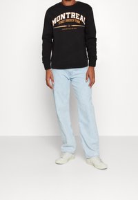 Sweatshirt nero con il logo "MONTREAL STREET HOCKEY TEAM" in arancione; abbinato a jeans azzurri chiari e sneakers color crema.