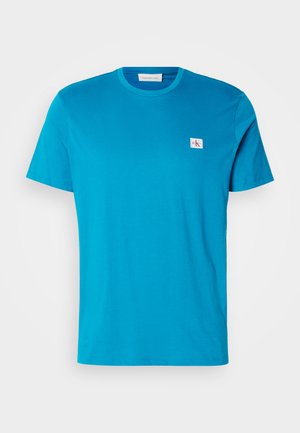 Turkis bomull t-skjorte med korte ermer, rund hals og en liten hvit logopatch på brystet. Glatt tekstur og avslappet passform.