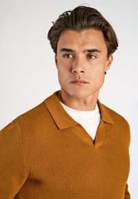 Polo shirt côtelé jaune moutarde avec un col en V et des manches courtes, superposé sur un t-shirt blanc. Présente une texture douce et un col bien défini.