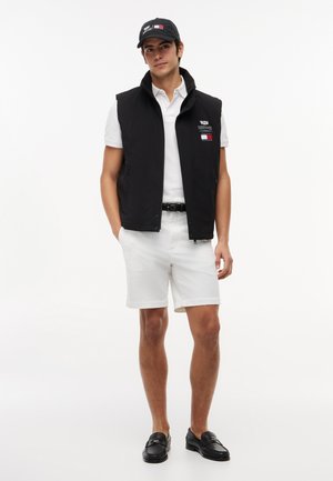 Jeune homme portant une casquette et un gilet noirs avec des logos, un polo blanc, un short blanc, une ceinture noire et des mocassins noirs, debout les mains dans les poches.