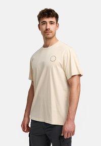 Camiseta de algodón beige con mangas cortas, cuello redondo y detalle gráfico circular en el pecho. Textura suave, diseño de corte recto.