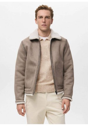 Giacca in finta pelliccia color cammello con collo in shearling bianco, chiusura con zip e polsini a costine. Indossata sopra un maglione lavorato a maglia beige e pantaloni chiari.
