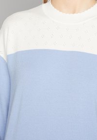 Ljusblå och vit tröja med en scalloped halsringning, med ett strukturerat mönster upptill och en slät design nedanför.