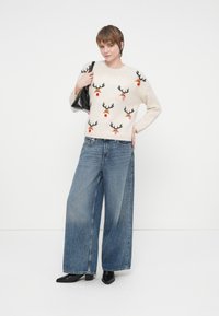 Sieviete, ģērbusies krēmkrāsas džemperī ar ziemeļbriežu sejām, ar platām zilām biksēm, melnām potīšu zābakiem, tur rokā melnu somu, stāv uz baltas fona.