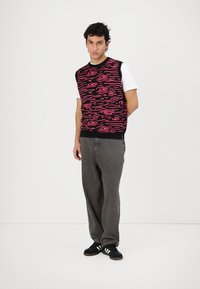Zwart mouwloos sweater met roze draaiende patronen, gedragen over een witte t-shirt. Gecombineerd met grijze broek en zwarte sneakers.