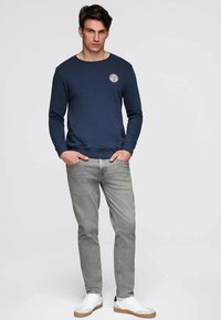 Marineblå sweatshirt med rund halsudskæring og frontpatch, kombineret med lysegrå jeans og hvide sneakers med tan detaljer. Glat stoftekstur.