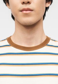 Νεαρό άτομο με κοντά σκούρα μαλλιά που φοράει λευκό πουκάμισο με οριζόντιες μπλε, καφέ και μουσταρδί ρίγες και καφέ γιακά.