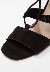 Sandale en suède noir avec un design à larges brides, bout ouvert et fermeture lacée à la cheville. Présente des coutures discrètes et des accents en métal.