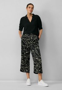 Blouse noire texturée avec un col en V et des manches retroussées, associée à un pantalon noir cropped avec un motif floral blanc et des baskets blanches.