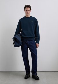 Sweat-shirt bleu marine avec un col rond et un petit logo doré, associé à un jean denim foncé et des chaussures noires, tenant un sac bleu.