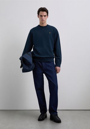 Navy blue sweatshirt with a round neckline and a small gold logo, paired with dark denim jeans and black shoes, holding a blue bag.