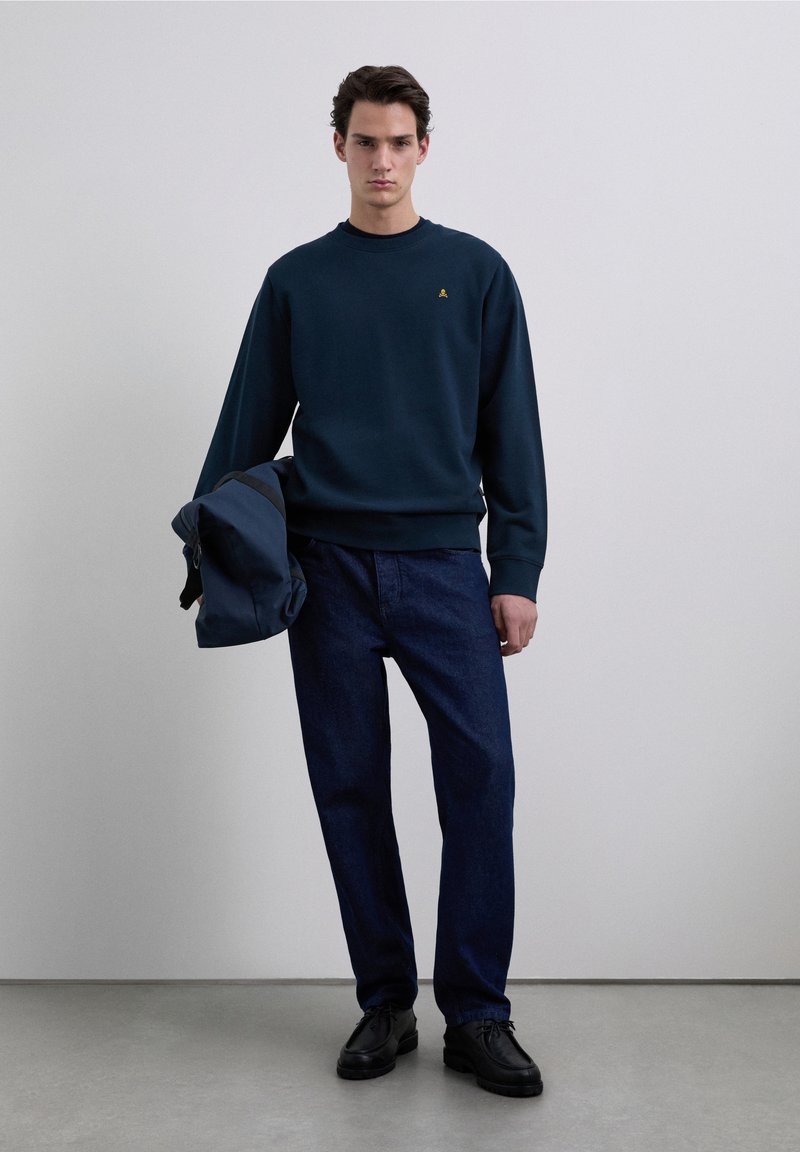 Sweat-shirt bleu marine avec un col rond et un petit logo doré, associé à un jean denim foncé et des chaussures noires, tenant un sac bleu.