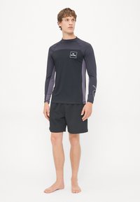 Czarny rash guard z długim rękawem i szarymi akcentami oraz logo Rip Curl, w parze z czarnymi, teksturowanymi szortami kąpielowymi, na modele bez butów.