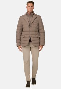 Beżowa kurtka puffer z pikowanym wzorem i stójką. Połączona z jasnobeżowymi spodniami i oliwkowymi trampkami.