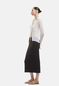 Blouse blanche et transparente à manches longues avec un détail de noeud à l'avant, associée à une jupe texturée brun foncé à longueur de cheville et des sandales plates à brides.