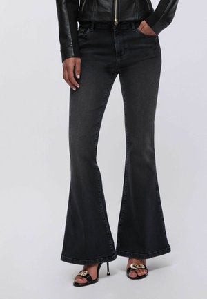 Žena u crnim širokim trapericama, crnoj kožnoj jakni i crnim sandalama s visokom petom s detaljem zlatnog lanca, stoji ispred jednobojne pozadine.