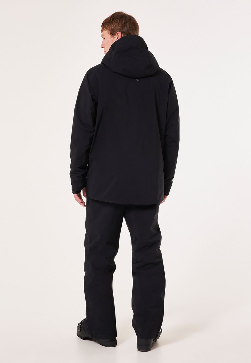 Schwarze wasserdichte Jacke mit Kapuze, kombiniert mit schwarzen Hosen. Die Merkmale umfassen lange Ärmel, eine lockere Passform und eine glatte Textur.