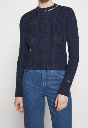 Persoon draagt een marineblauwe kabelgebreide trui met "Tommy Jeans" op de kraag en blauwe jeans met hoge taille, staand tegen een effen achtergrond.