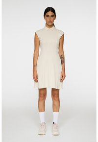 Nainen, jolla on tumma tukka, pukeutuneena beigeen hihattomaan kauluspaitamekkoon, valkoisiin sukkiin ja beigeihin tennareihin, seisoo yksinkertaista valkoista taustaa vasten.