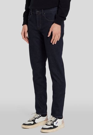 Jean en denim bleu foncé avec une coupe slim, présentant des poches avant, un zip à bouton et une couture contrastante. Porté avec des baskets noir et blanc.