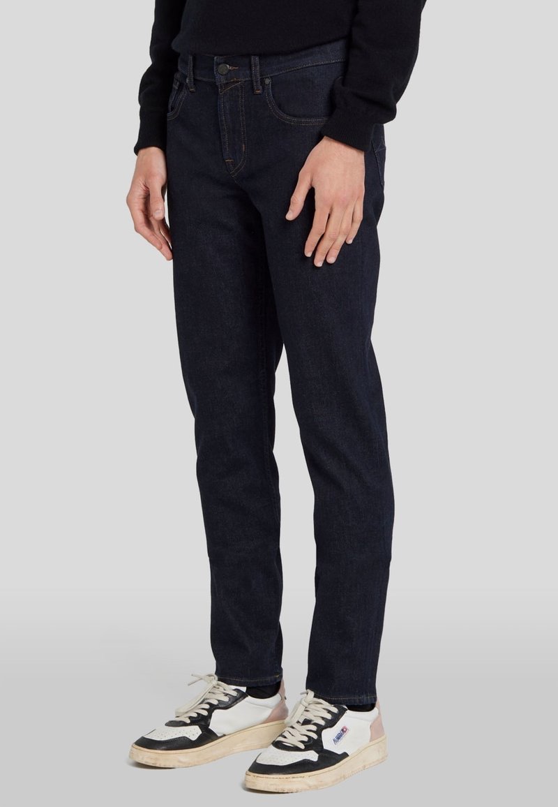 Donkerblauwe denim jeans met een slim fit, voorzien van voorzakken, een knoopsluiting en contrasterende stiksels. Gecombineerd met zwart-witte sneakers.