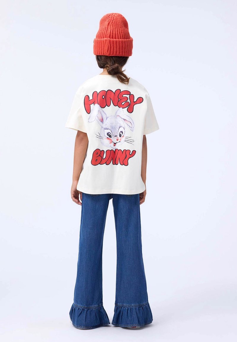 Weißes Kurzarm-T-Shirt mit einem Karikaturhase-Graphic, kombiniert mit blauen Schlagjeans mit gerafften Säumen. Das Modell trägt eine rote Strickmütze.