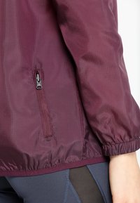 Chaqueta ligera color burdeos con un acabado texturizado, que presenta un bolsillo con cremallera y puños elásticos para un ajuste seguro. Líneas de diseño acentuadas visibles.