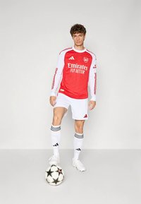Rød og hvid langærmet fodboldtrøje med "Emirates Fly Better"-logo, kombineret med hvide shorts og sokker med sorte striber, stående ved en fodbold.